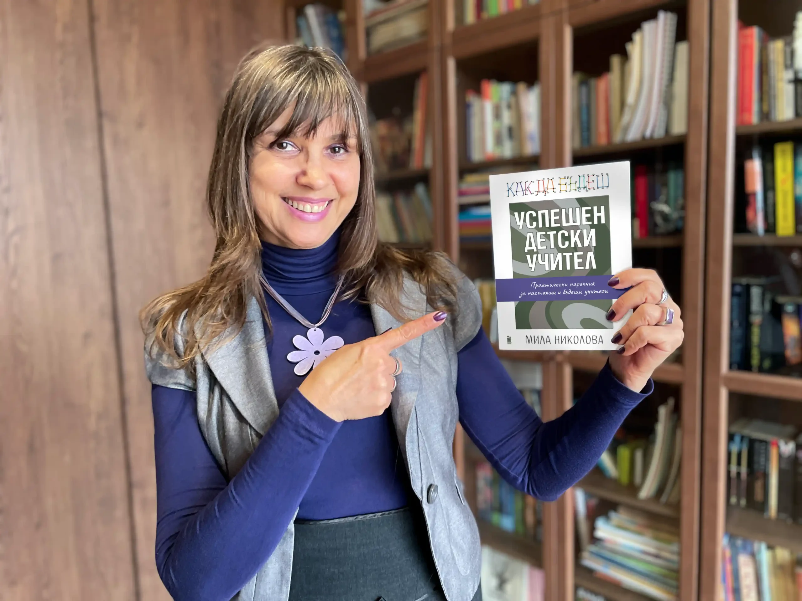 Мила държи наръчника „Как да бъдеш успешен детски учител“ в ръка, сочи го с пръст и се усмихва.