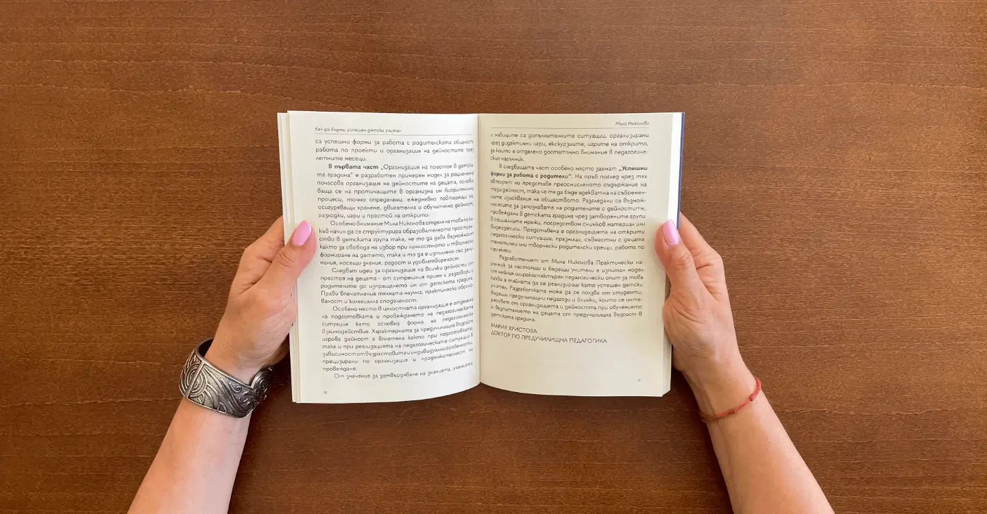 Приближен кадър на две ръце, облегнати върху маса, държащи отворена книга.