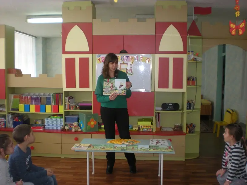 Мила Николова държи книжка и говори на деца в детска градина. Вижда се интериора и преградна стена във формата на замък.